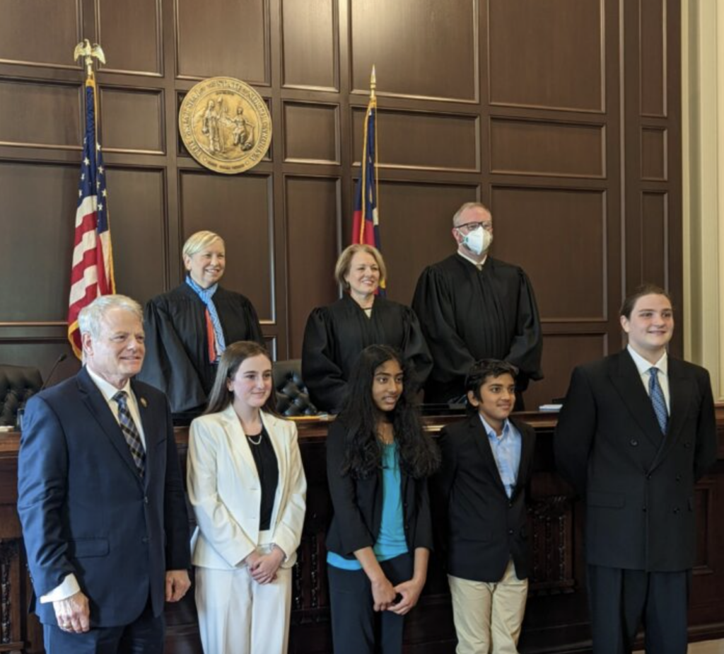 8 people standing in a court room in front of a dark wooden wall with decorative golden emblem and the American and NC flags