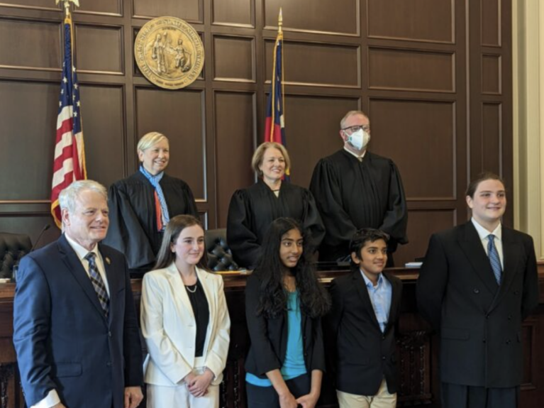 8 people standing in a court room in front of a dark wooden wall with decorative golden emblem and the American and NC flags
